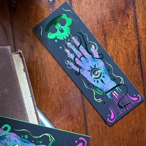 May include: A black bookmark featuring a green skull and a hand with an eye in the palm. The hand is surrounded by swirling lines and dots in shades of blue, purple, and pink. The bookmark is lying on a wooden surface.