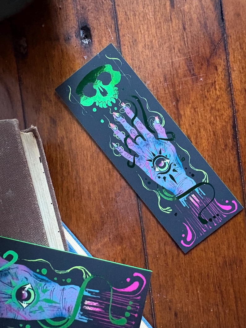 May include: A black bookmark featuring a green skull and a hand with an eye in the palm. The hand is surrounded by swirling lines and dots in shades of blue, purple, and pink. The bookmark is lying on a wooden surface.