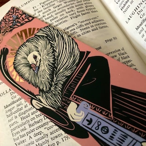 May include: A pink bookmark with a black and white illustration of a bird with a long neck and a long beak. The bird is holding a blue and white object in its hand. The bookmark has the text "COLLAR DU" and "Ge" on it.