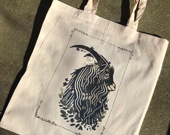May include: A white tote bag with a black and white illustration of a goat's head with floral accents. The goat is surrounded by a black frame with a floral border.