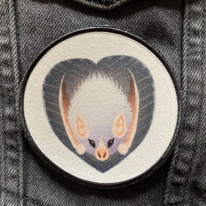 May include: A white bat with pink ears and a black heart-shaped background. The bat is depicted in a cartoon style with large eyes and a small nose. The heart is outlined in black and has a white background.