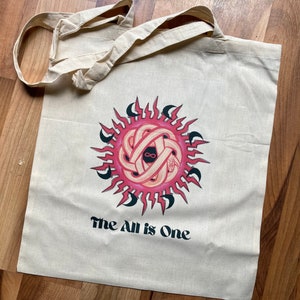 May include: A white canvas tote bag with a pink and black graphic of a sun with an infinity symbol inside. The text "The All is One" is printed below the graphic.