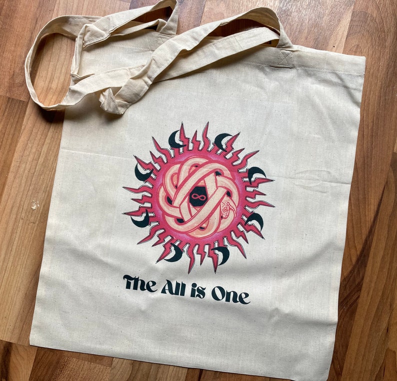 May include: A white canvas tote bag with a pink and black graphic of a sun with an infinity symbol inside. The text "The All is One" is printed below the graphic.