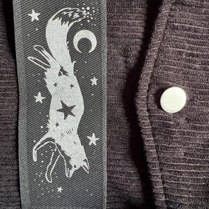 May include: A black fabric patch with a white illustration of a fox, stars, and a crescent moon. The patch is sewn onto a dark, textured fabric, with a silver button visible.