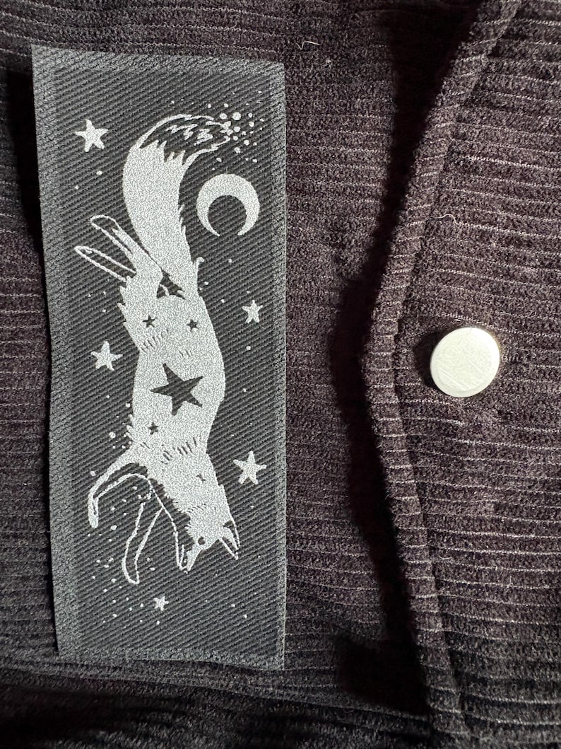 May include: A black fabric patch with a white illustration of a fox, stars, and a crescent moon. The patch is sewn onto a dark, textured fabric, with a silver button visible.