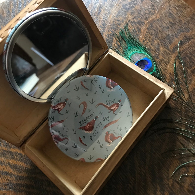 May include: A round, blue compact mirror with a design of pheasants and the words "Strut Pretty Boy" and "Babaam!" on a white background.