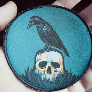 May include: A black crow perched on a white skull with a teal background. The crow is facing left and the skull is surrounded by dark green foliage.