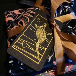 May include: A stack of black fabric with a gold foil design is wrapped in a gold ribbon and tied with a bow. A black card with a gold foil design of a creature with a tail is attached to the ribbon.