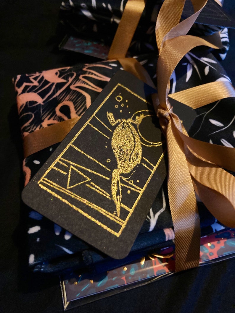 May include: A stack of black fabric with a gold foil design is wrapped in a gold ribbon and tied with a bow. A black card with a gold foil design of a creature with a tail is attached to the ribbon.