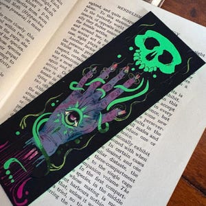 May include: Black bookmark with a neon green skull and hand illustration. The hand has an eye in the palm and long fingernails. Green, vine-like lines connect the skull and hand. The bookmark is on top of an open book.