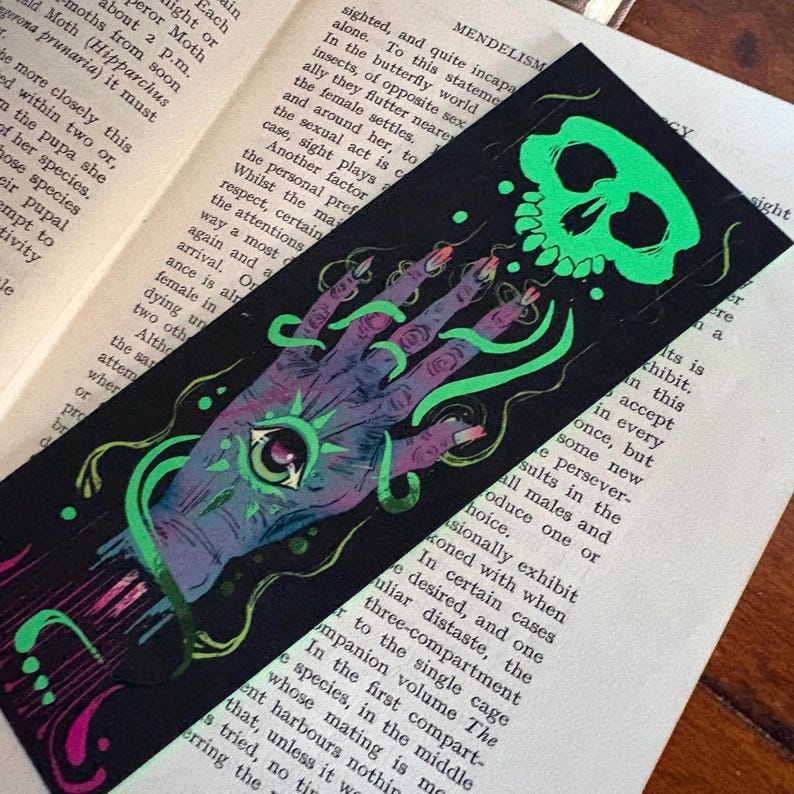 May include: Black bookmark with a neon green skull and hand illustration. The hand has an eye in the palm and long fingernails. Green, vine-like lines connect the skull and hand. The bookmark is on top of an open book.