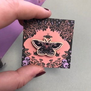 May include: A black and white wooden moth pin with a skull design on its body. The pin is on a pink background with a black and white floral design and skulls.