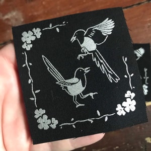 May include: Black fabric patch with a white outline of two magpies and floral designs. The magpies are facing opposite directions, one is flying and the other is perched on the ground.