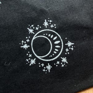 May include: A white crescent moon and sun design with a circle of white stars on a black background.