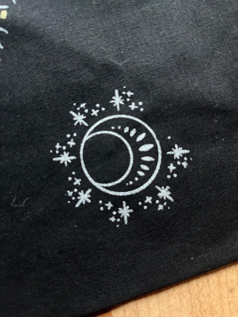 May include: A white crescent moon and sun design with a circle of white stars on a black background.