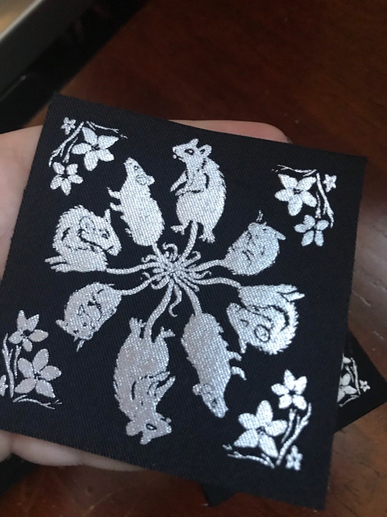 May include: A black fabric patch with a silver design of seven mice in a circle with their tails intertwined. The mice are surrounded by small silver flowers.