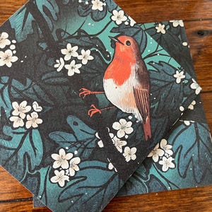 May include: A robin perched on a branch with white flowers, surrounded by dark green leaves. The bird is red and white with a black eye and a long, pointed beak. The background is a dark green and black pattern.