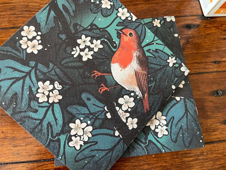 May include: A robin perched on a branch with white flowers, surrounded by dark green leaves. The bird is red and white with a black eye and a long, pointed beak. The background is a dark green and black pattern.