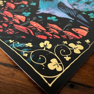 Puede incluir: Una tarjeta negra con un borde dorado y una ilustraci&oacute;n caprichosa de un p&aacute;jaro azul con las alas extendidas, rodeado de setas rojas y naranjas y tr&eacute;boles dorados sobre un fondo azul oscuro.