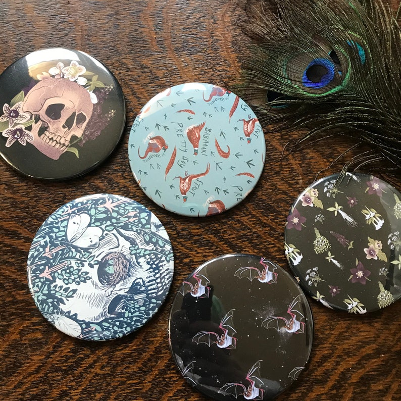 May include: Set of six round compact mirrors with various designs. The mirrors have a black background with colorful designs. One mirror features a skull with flowers, another has a pattern of birds and arrows, another has a pattern of bats, another has a pattern of flowers, and another has a pattern of leaves and flowers.
