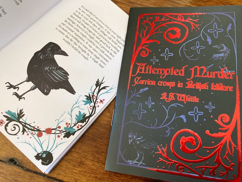 May include: A black book cover with a red and blue design featuring a crow. The title of the book is "Attempted Murder: Carrion Crows in British Folklore" by R.S. Whittle.