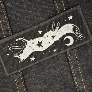 May include: A rectangular black patch featuring a white illustration of a fox leaping among stars and a crescent moon. The patch is sewn onto a dark denim fabric with visible stitching.