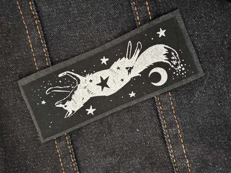 May include: A rectangular black patch featuring a white illustration of a fox leaping among stars and a crescent moon. The patch is sewn onto a dark denim fabric with visible stitching.