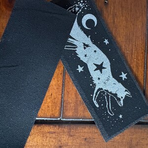 May include: Two black fabric patches. One patch features a silver fox design with a crescent moon, stars, and a large star. The other patch is a solid black rectangle. The patches are on a wooden surface.