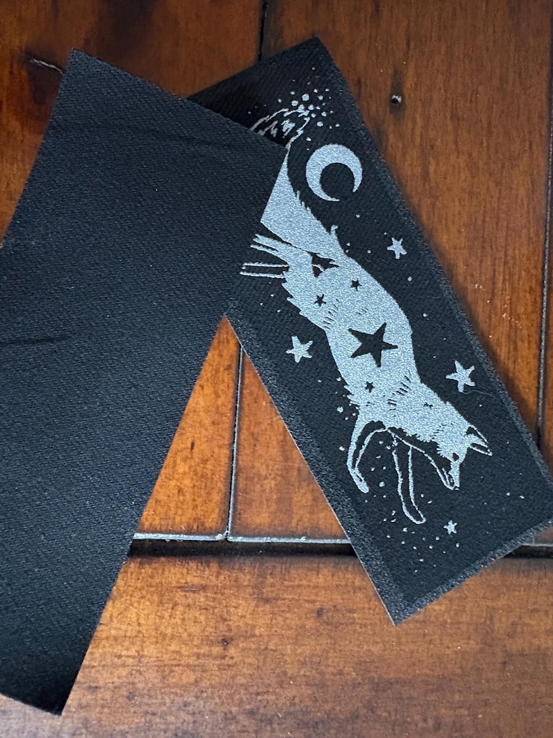 May include: Two black fabric patches. One patch features a silver fox design with a crescent moon, stars, and a large star. The other patch is a solid black rectangle. The patches are on a wooden surface.