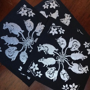 May include: Three black fabric squares with white embroidered designs of mice and flowers. The mice are arranged in a circle around a central point, with their tails extending outwards. The flowers are scattered around the edges of the squares.