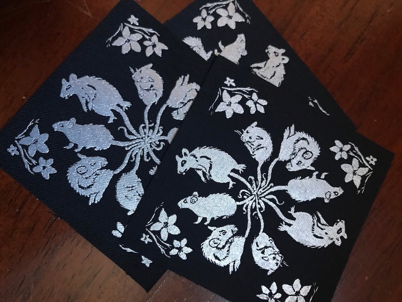 May include: Three black fabric squares with white embroidered designs of mice and flowers. The mice are arranged in a circle around a central point, with their tails extending outwards. The flowers are scattered around the edges of the squares.