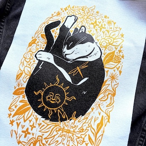 May include: A black and white cat illustration with a smiling sun and dragonfly design. The cat is surrounded by a gold floral pattern. The artwork is printed on a white fabric.