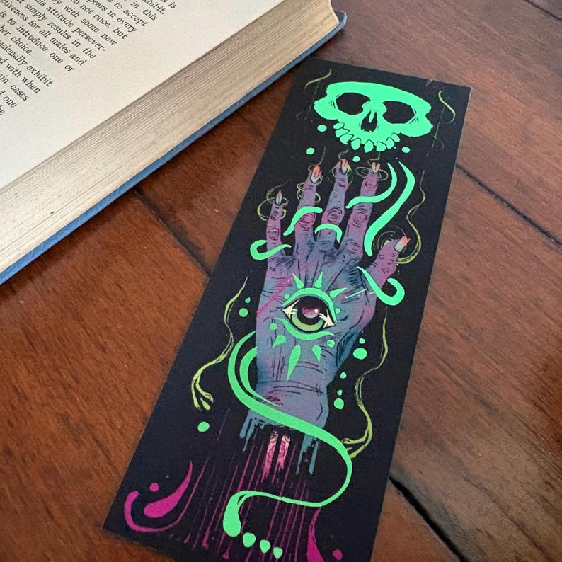 May include: A black bookmark with a detailed illustration. It features a purple hand with an eye in the palm, a green skull, and green swirling lines. The bookmark is resting on a wooden surface next to a book.