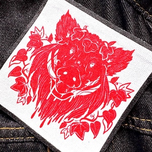 May include: Square patch with a red illustration of a pig's head adorned with leaves and flowers. The patch has a white background and a black border. The patch is on a dark denim fabric.