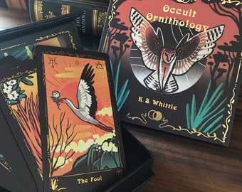 May include: A deck of tarot cards with a black background and colourful illustrations. The card shown is "The Fool" with a white bird flying in a sunset sky. The box features a black and gold design with the title "Occult Ornithology" and the name "K.S. Whittle".