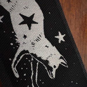 May include: Black patch featuring a stylized silver fox design with stars. The fox is depicted with a celestial theme, surrounded by various sized stars. The patch has a textured appearance, with a detailed illustration of the fox and stars.