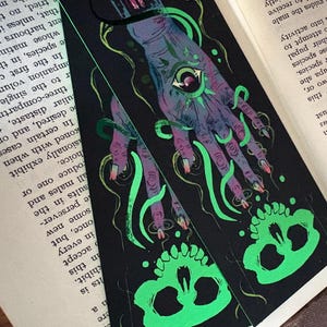 May include: Black bookmark with a detailed illustration of a hand with an eye in the palm, green tentacles, and a skull. The hand is purple and the skull is neon green. The bookmark is on top of an open book.
