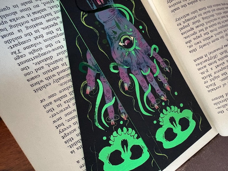 May include: Black bookmark with a detailed illustration of a hand with an eye in the palm, green tentacles, and a skull. The hand is purple and the skull is neon green. The bookmark is on top of an open book.