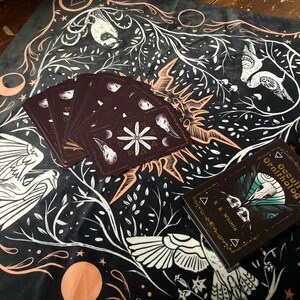 May include: A black velvet cloth with a white and gold design featuring birds, moons, and stars. The cloth has a deck of cards and a book titled "Occult Ornithology" on top of it.