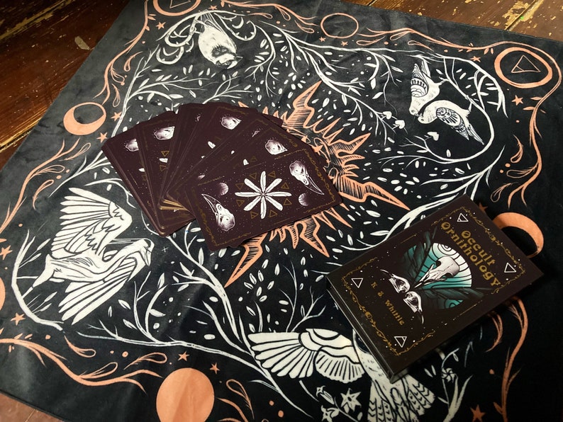 May include: A black velvet cloth with a white and gold design featuring birds, moons, and stars. The cloth has a deck of cards and a book titled "Occult Ornithology" on top of it.