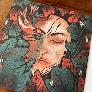 May include: A close-up of a book cover featuring a woman's face surrounded by green and red leaves, with a small bird perched on a branch. The woman's eyes are closed and she appears to be sleeping. The text on the page reads "By boo that the v Robin their as Glasgo the 6th times th In many visiting"