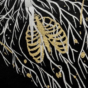 May include: A black fabric background with a white and gold design. The design features a rib cage with gold glitter and white branches.