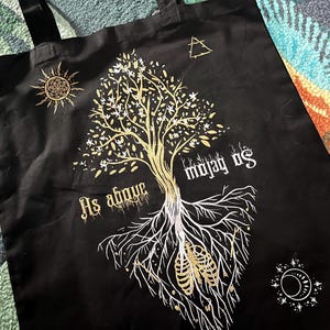 May include: Black tote bag with a white and gold design of a tree with roots and the text "As above so below".