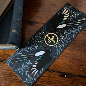 Puede incluir: Un marcapáginas en blanco y negro con un diseño dorado que presenta dos urracas con detalles dorados sobre un fondo verde oscuro con puntos dorados y hojas.