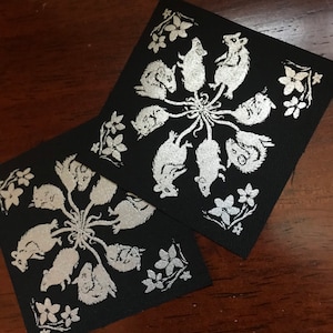 May include: Two black fabric squares with a silver design of mice in a circle. The mice are facing inward and have their tails intertwined. There are small silver flowers around the mice.