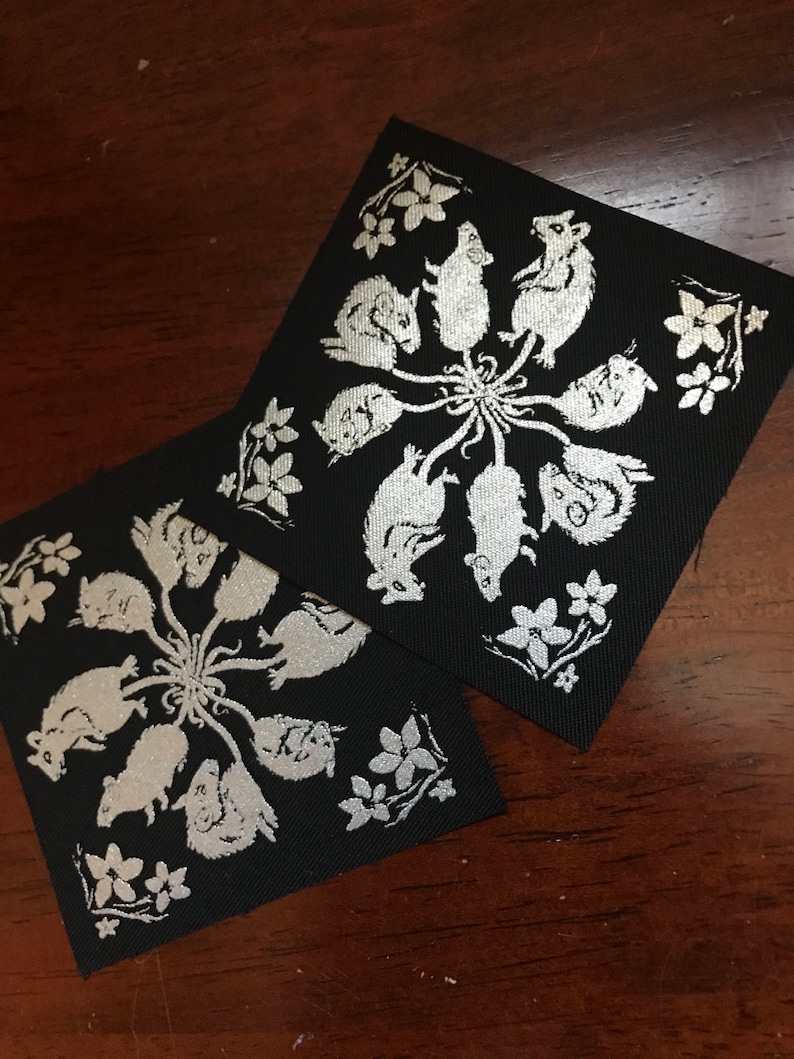 May include: Two black fabric squares with a silver design of mice in a circle. The mice are facing inward and have their tails intertwined. There are small silver flowers around the mice.