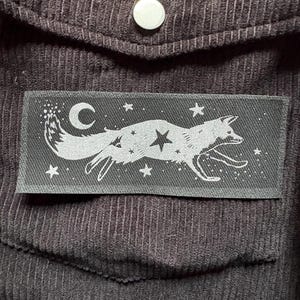 May include: A dark gray rectangular patch featuring a running fox design with a crescent moon, stars, and a central star. The patch is sewn onto a dark brown corduroy fabric, with a pocket and a button visible.
