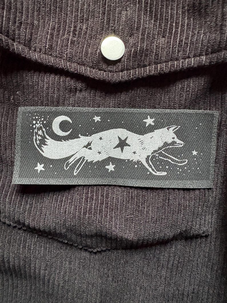 May include: A dark gray rectangular patch featuring a running fox design with a crescent moon, stars, and a central star. The patch is sewn onto a dark brown corduroy fabric, with a pocket and a button visible.