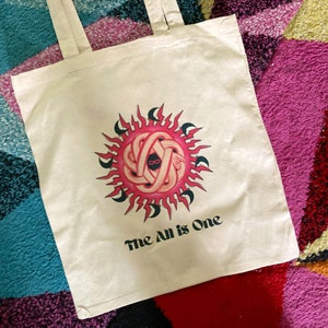 May include: A white tote bag with a pink and black sun design and the text "The All is One".
