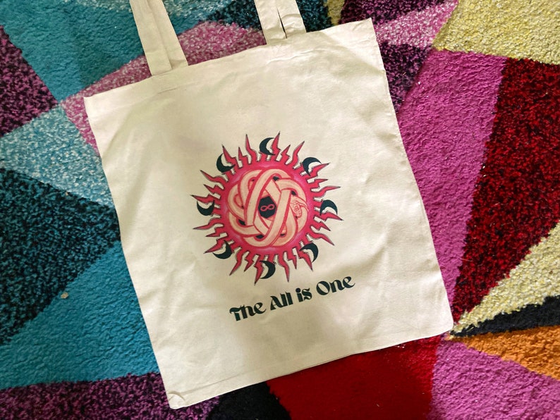 May include: A white tote bag with a pink and black sun design and the text "The All is One".